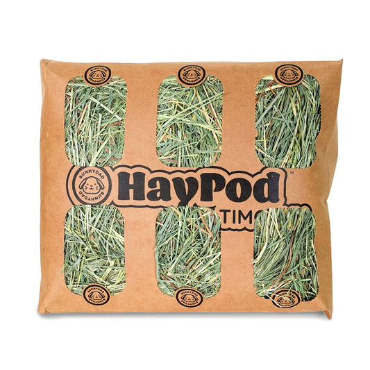 Opened on a white background, the brown paper BunnyHay Pod reveals fresh green hay inside. In the center of the Hay Pod, there is the BunnyDad logo and the text 'HayPod Timothy.'